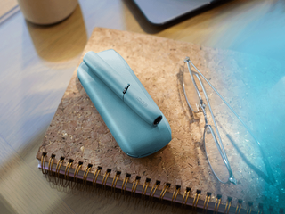 Turquoise IQOS Originals Duo device on a notebook next to glasses.