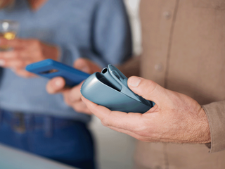 Hands holding a turquoise IQOS Originals device and a mobile phone.