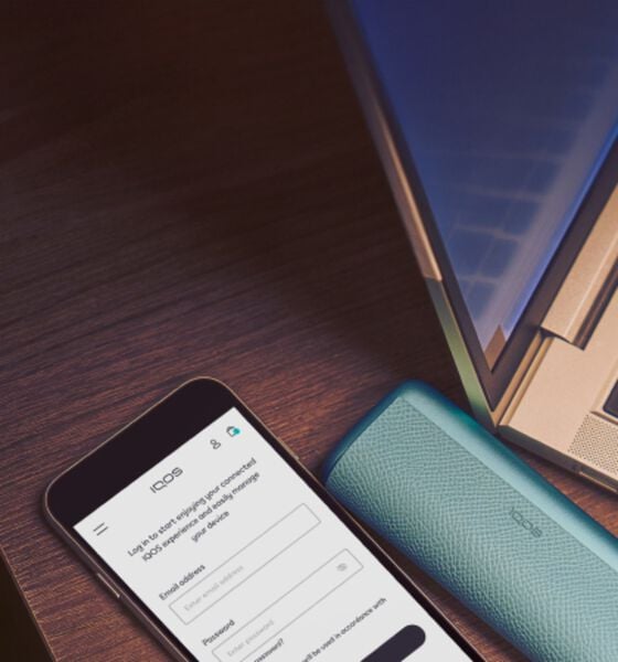IQOS ILUMA i Prime in teal placed next to a smartphone showing the IQOS app login screen and a laptop on a wooden desk.