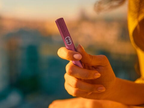 شخص يحمل جهاز IQOS ILUMA i Prime باللون البرقوقي المعدني مع خلفية مدينة ضبابية عند غروب الشمس.