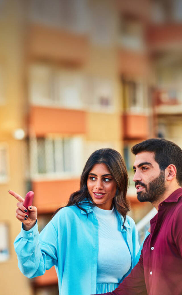 امرأة ترتدي زيًا أزرق فاتح تشير إلى شيء ما بينما تمسك جهاز IQOS، وتقف بجوار رجل يرتدي قميصًا بلون أحمر داكن. هما في مكان خارجي في بيئة حضرية مع مبانٍ ضبابية في الخلفية.