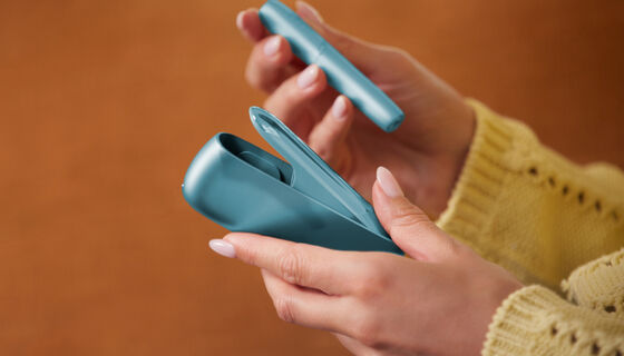 Hands holding IQOS Originals Duo heated tobacco holder and pocket charger in turquoise color.