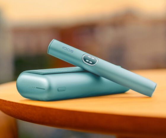 IQOS ILUMA i Prime in teal with holder and charger placed on a wooden table with a blurred background.
