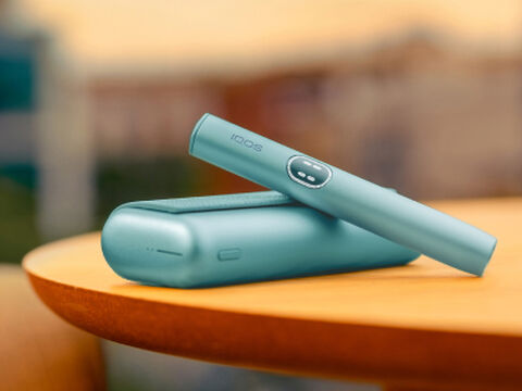 جهاز IQOS ILUMA i Prime باللون الأزرق المعدني مع الحامل والشاحن، موضوع على طاولة خشبية بخلفية ضبابية
