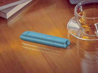 جهاز IQOS Originals One بلون التركواز موضوع على مكتب بجانب فنجان شاي.