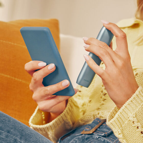 Hands holding a mobile phone and IQOS Originals One device, downloading the app to get a full professionnal maintenance.