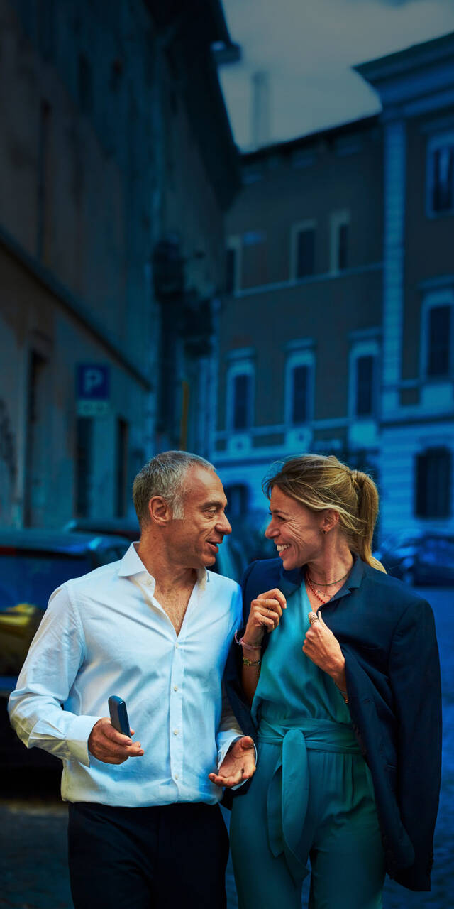 A man and a woman walking together, smiling and having a conversation. The man is holding a phone, and both are dressed casually, enjoying their time outside in a vibrant urban setting at dusk. The atmosphere is warm and friendly.