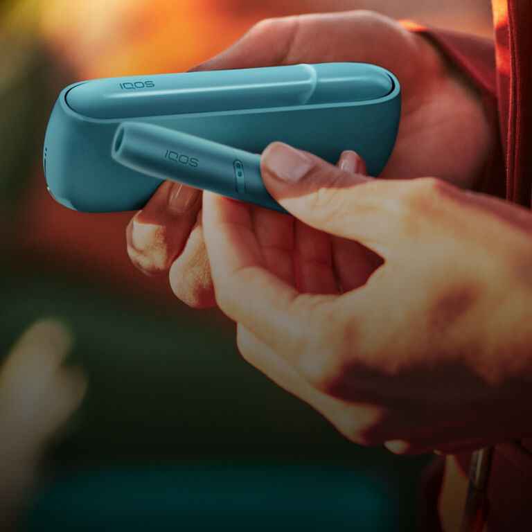 يد تمسك أنبوب تسخين وشاحن جيب جهاز IQOS Originals Duo بلون التركواز.
