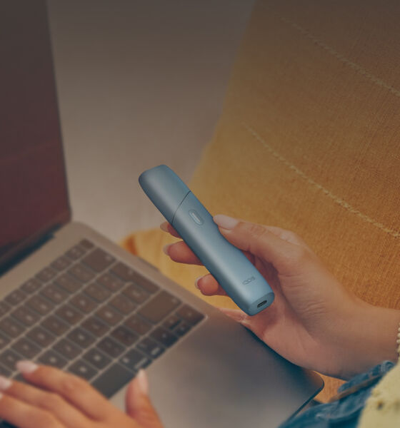 Woman holding an IQOS Originals ONE heated tobacco device and typing on laptop