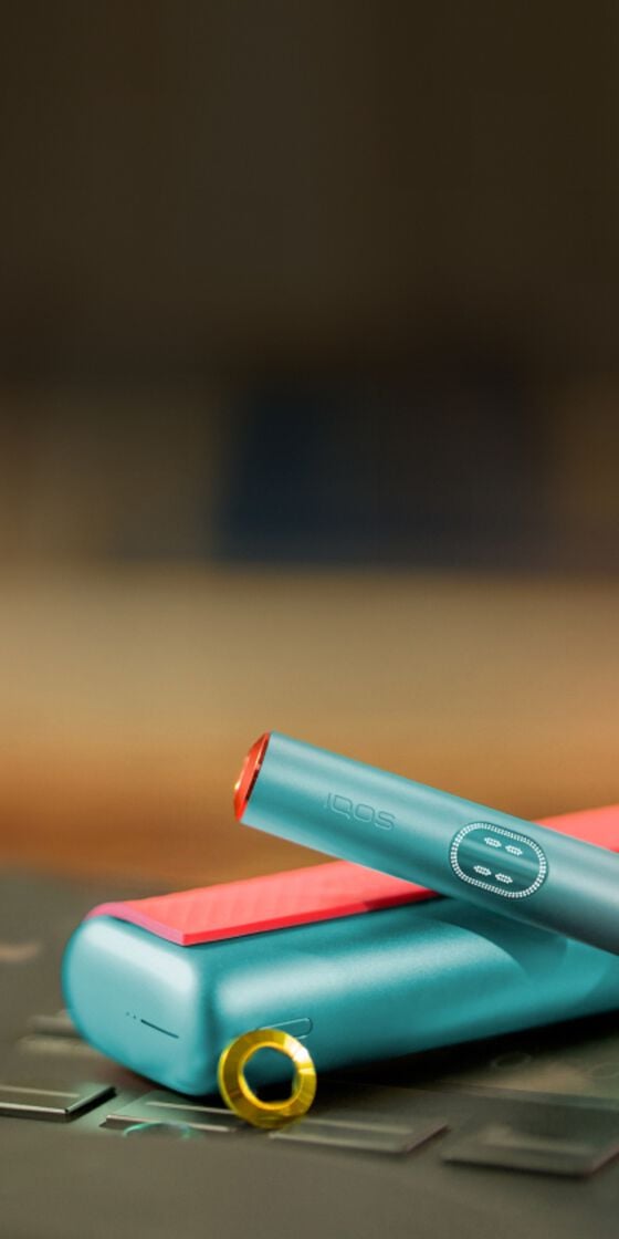 جهاز IQOS ILUMA i Prime باللون الأزرق المعدني مع لمسات حمراء، مع الحامل والشاحن على خلفية ضبابية.